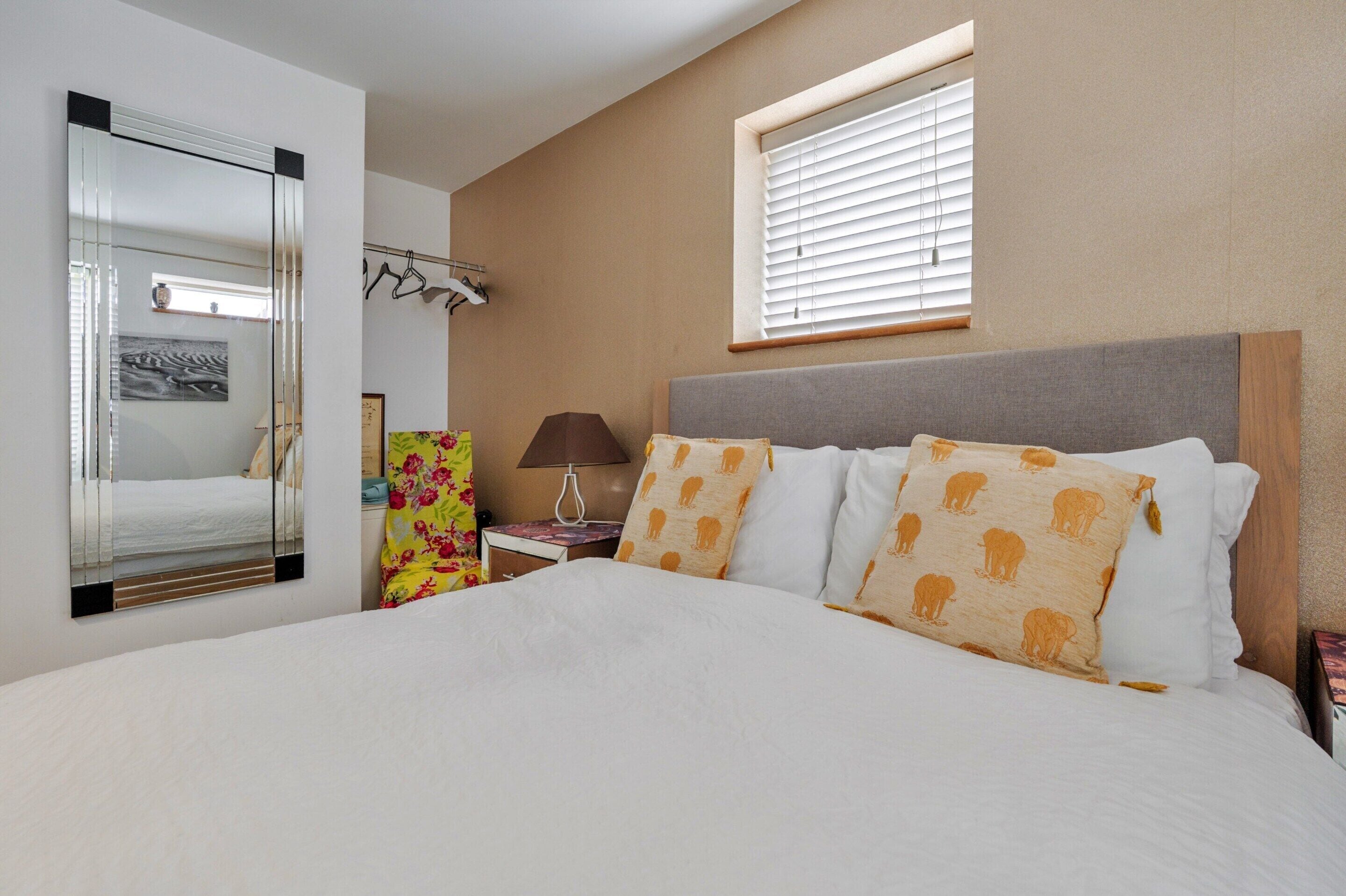 A hotel room with a large bed, a bedside table with a lamp, a window with blinds, and a mirror on the wall.