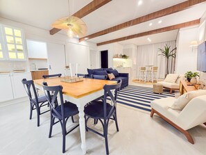 A modern living room with a dining area, featuring a wooden dining table, dark chairs, a blue sofa, and a white armchair.