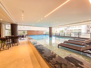 An indoor swimming pool with lounge chairs and a bar area. - Bravavel na Praia Brava by LikeHome (Balneário Camboriú)