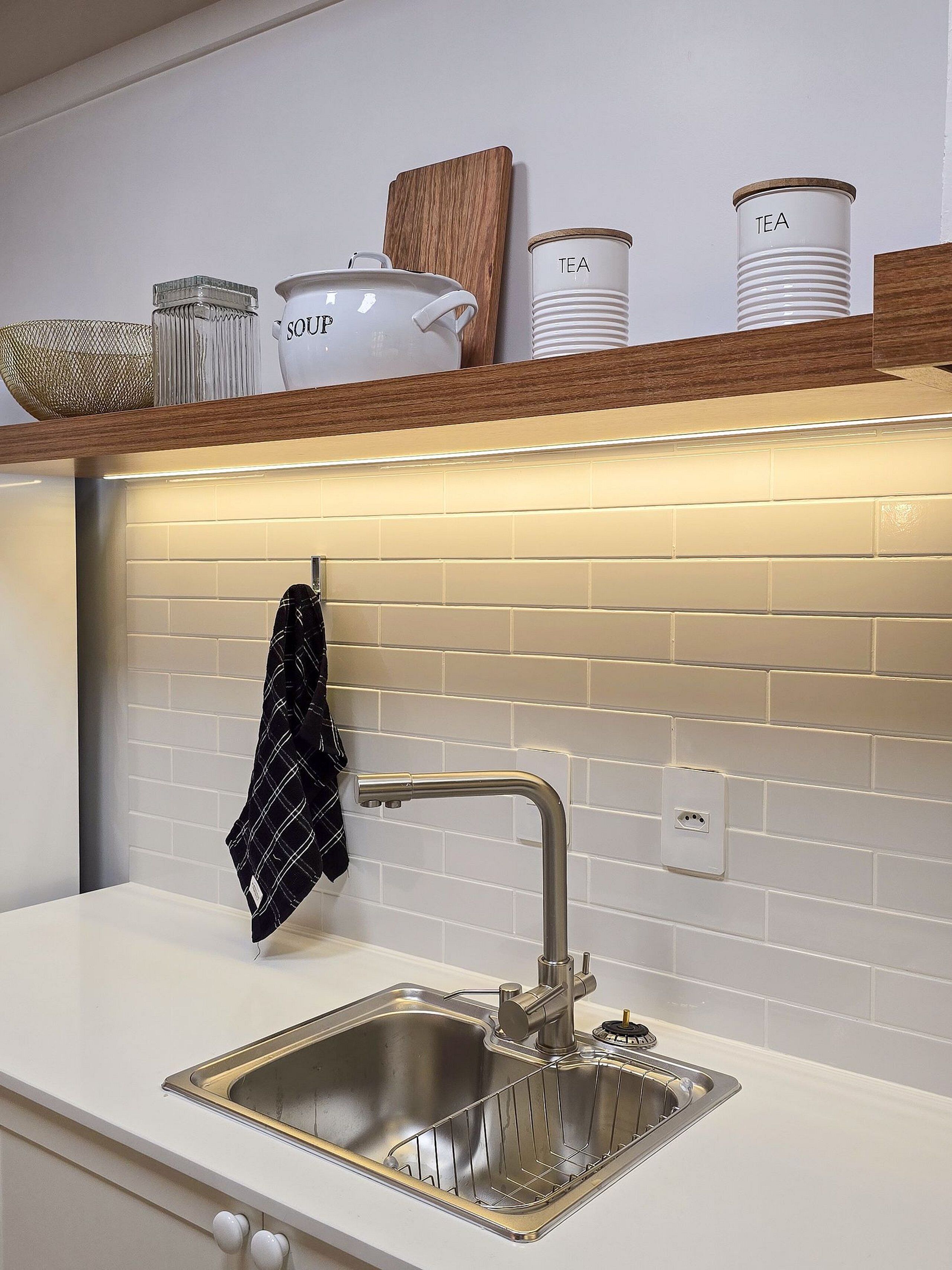 Eine moderne Küche mit Spüle aus Edelstahl, einem Holzregal darüber und einer gefliesten Spritzschutzwand.