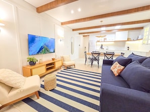 Uma sala moderna com televisão de tela plana, um sofá, uma área de jantar com cadeiras e uma cozinha ao fundo.