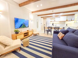 Uma sala moderna com televisão de tela plana, um sofá, uma área de jantar com cadeiras e uma cozinha ao fundo.
