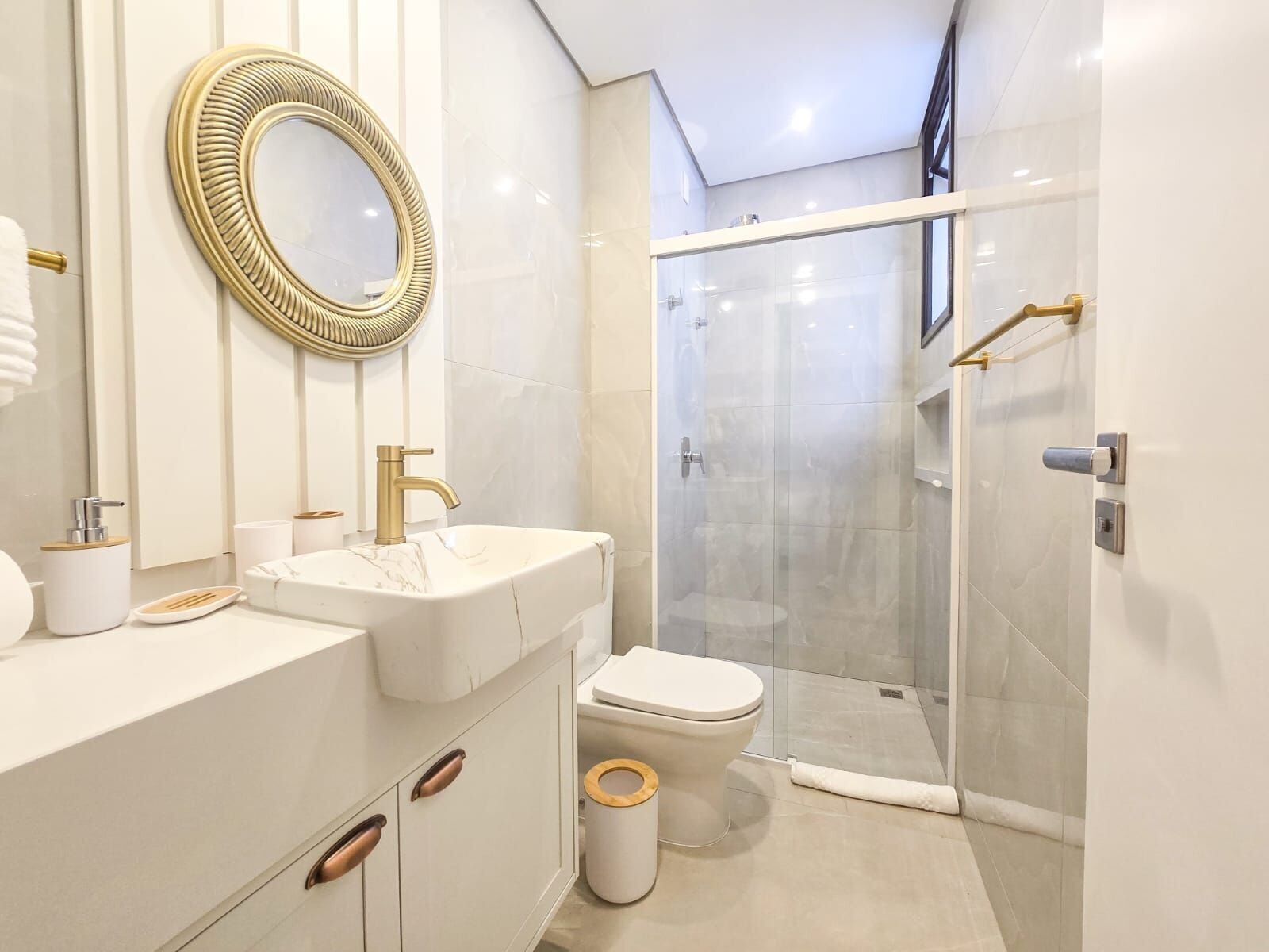 A modern bathroom with a white vanity, gold faucets, a round mirror, a glass-enclosed shower, and a toilet with a wooden bidet seat.