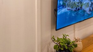 Un televisor de pantalla plana montado en la pared, una lámpara de pared y un estante de madera con una planta en maceta y un objeto decorativo.