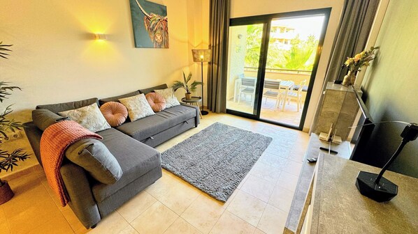 A modern living room with a grey sofa, a grey rug, and a large window with a view of outdoor furniture.