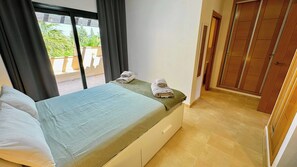 Una camera d'albergo con un letto, una finestra con tende e vista sul verde circostante, più una porta in legno.