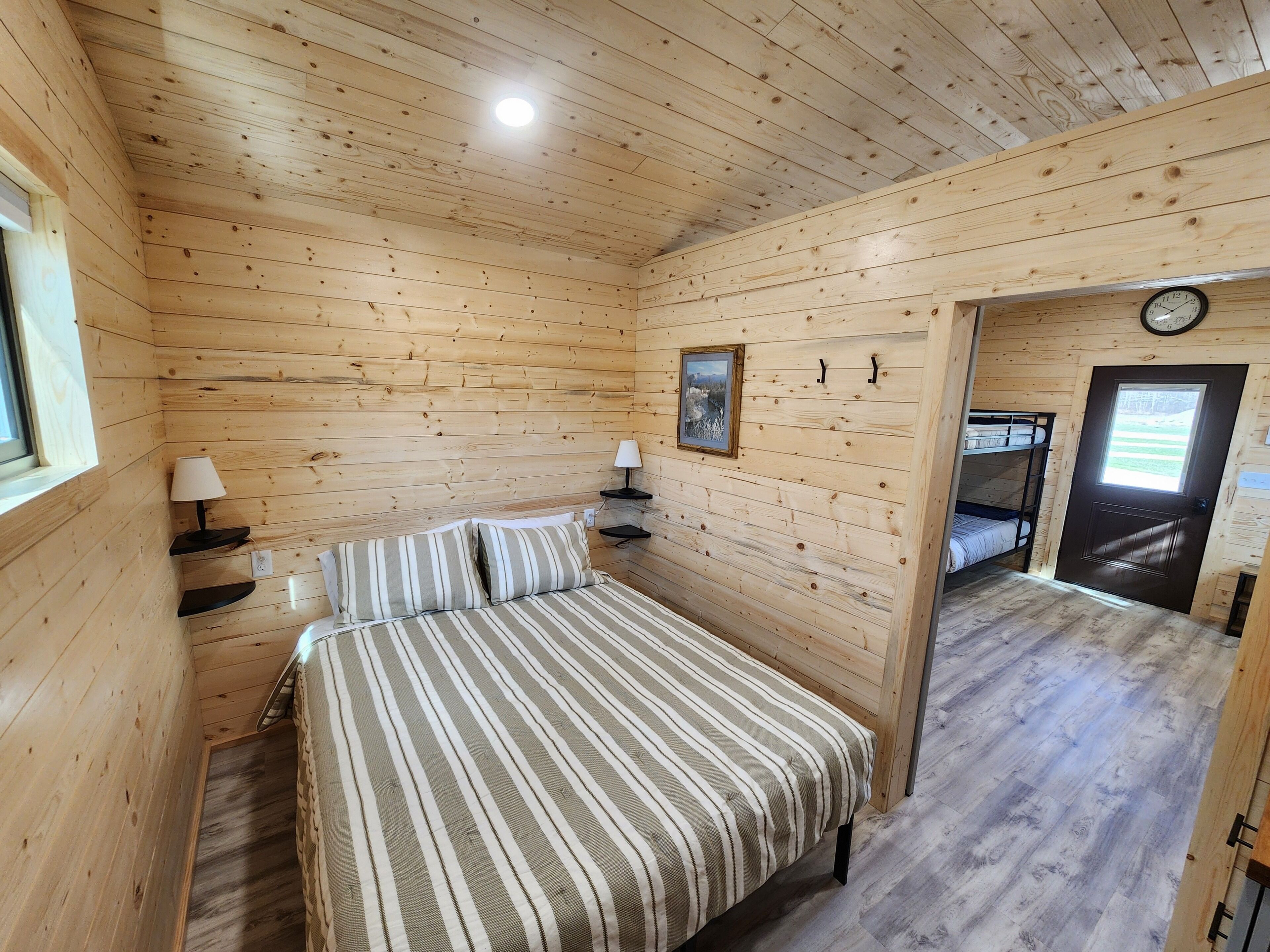 A wooden cabin room with a bed, bedside lamps, a framed picture, a clock, and a bunk bed in the adjacent room.