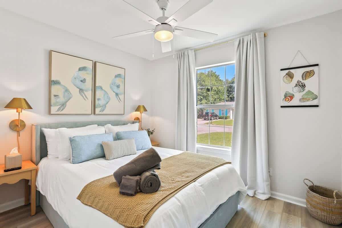 Une chambre à coucher comprenant un lit, deux lampes fixées au mur, un ventilateur de plafond, une fenêtre avec des rideaux et des œuvres d’art accrochées aux murs.