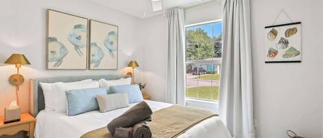 Une chambre à coucher comprenant un lit, deux lampes fixées au mur, un ventilateur de plafond, une fenêtre avec des rideaux et des œuvres d’art accrochées aux murs.