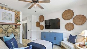 A living room with a stone fireplace, a ceiling fan, a flat-screen TV, and a blue cabinet. - Coastal Comfort - 15 min to Beach, 10 min to City (Savannah)