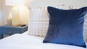 A bed with white bedding, a dark blue pillow, and a striped pillow. There is a bedside table with a lamp and a remote control. - Coastal Comfort - 15 min to Beach, 10 min to City (Savannah)