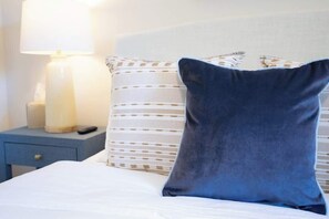 A bed with white bedding, a dark blue pillow, and a striped pillow. There is a bedside table with a lamp and a remote control. - Coastal Comfort - 15 min to Beach, 10 min to City (Savannah)