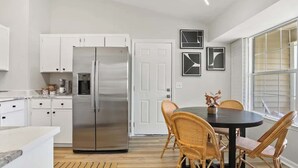Eine moderne Küche mit weißen Schränken, einem Edelstahlkühlschrank, einem Esstisch mit Korbstühlen und gerahmten Kunstwerken an der Wand.