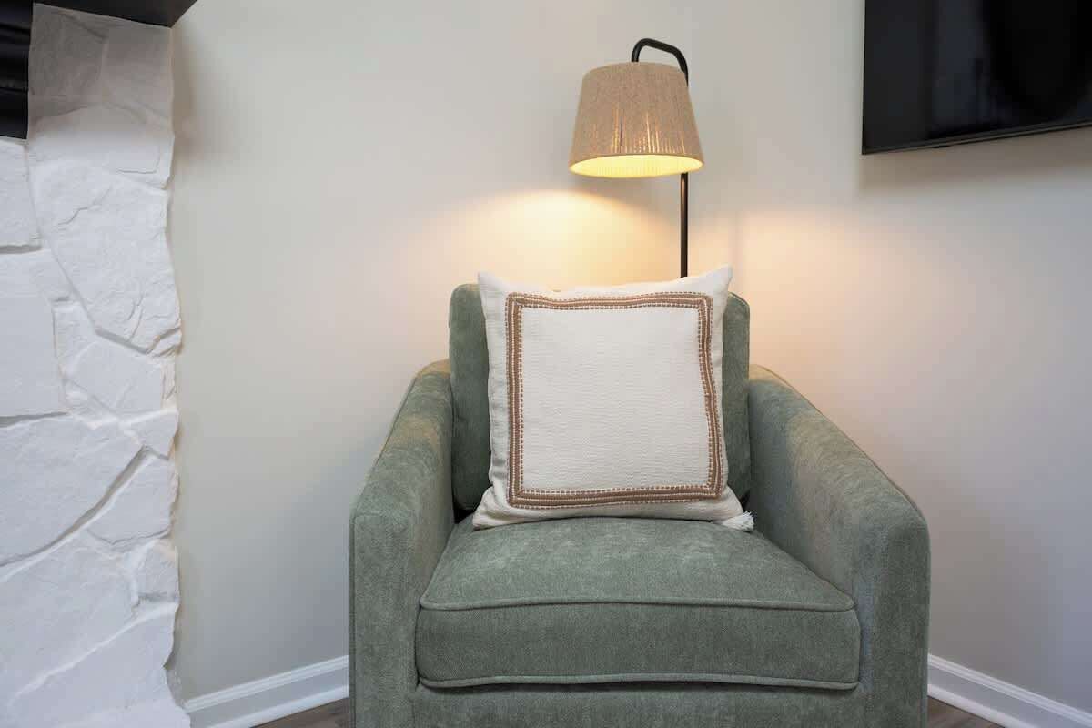 Un fauteuil vert avec un coussin blanc, placé devant un mur uni, avec une lampe et un téléviseur à écran plat fixé au-dessus.
