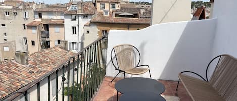 A balcony with two wicker chairs and a small table, overlooking a cityscape with terracotta rooftops.