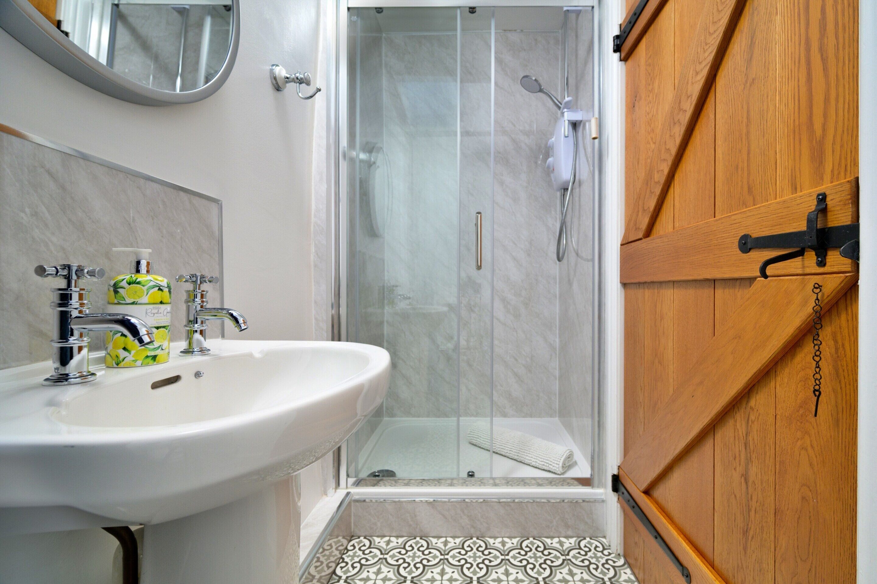 A bathroom with a white sink, a mirror, a shower enclosure, and a wooden door.