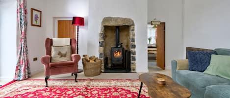 A cozy living room with a red patterned carpet, a plush armchair, a blue sofa, a wooden coffee table, and a stone fireplace with a wood-burning stove.