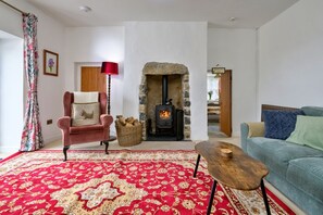 A cozy living room with a red patterned carpet, a plush armchair, a blue sofa, a wooden coffee table, and a stone fireplace with a wood-burning stove. - Scenic Snowdonia Border Cottage for 4 (Caernarfon)