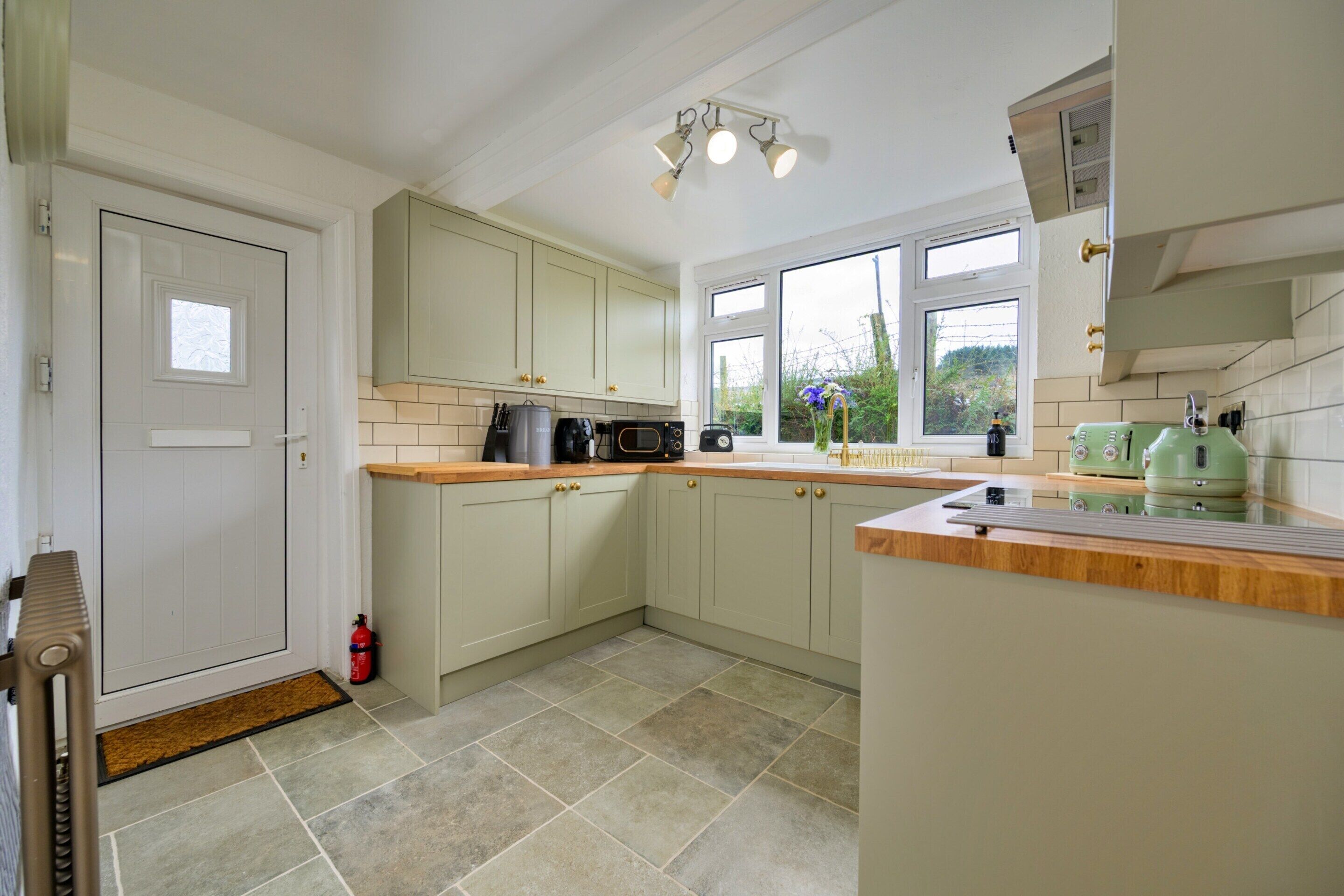 A kitchen with white cabinets, a microwave, a kettle, and a window with a view of greenery.