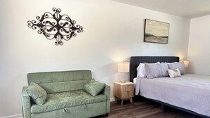 Ein Schlafzimmer mit einem grünen Sofa, einem Bett mit vielen Kissen, einem Nachttisch mit Lampe, Holzboden und einem Wandbild.