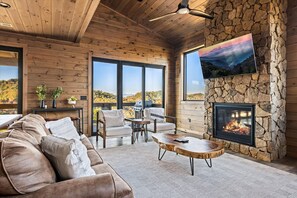 Ein gemütliches Wohnzimmer mit Kamin aus Stein, Holzwänden und großen Fenstern mit Blick auf eine reizvolle Landschaft.
