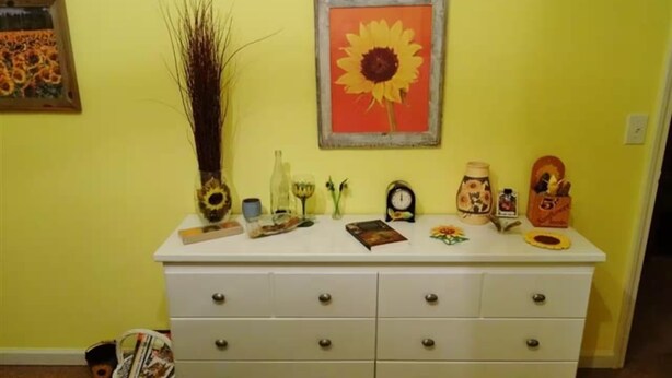 A room with a white dresser against a yellow wall, featuring a sunflower painting, a vase, a clock, and various decorative items. - Nature Lover’s Retreat: Cozy 2-Bedroom Pine Mountain Cabin near Callaway Gardens (Pine Mountain)