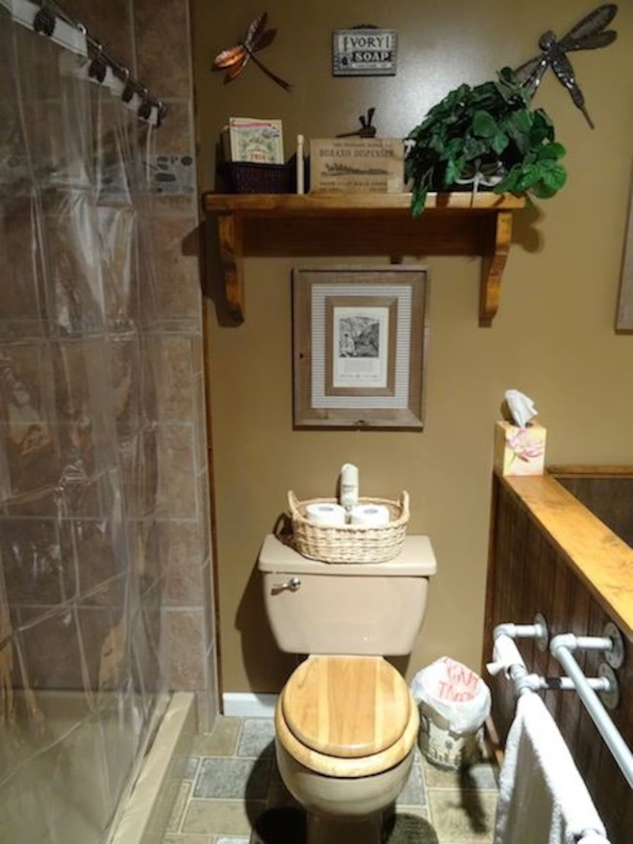 A bathroom with a toilet, a wooden bath mat, a shower curtain, a shelf with toiletries, and a framed picture on the wall.