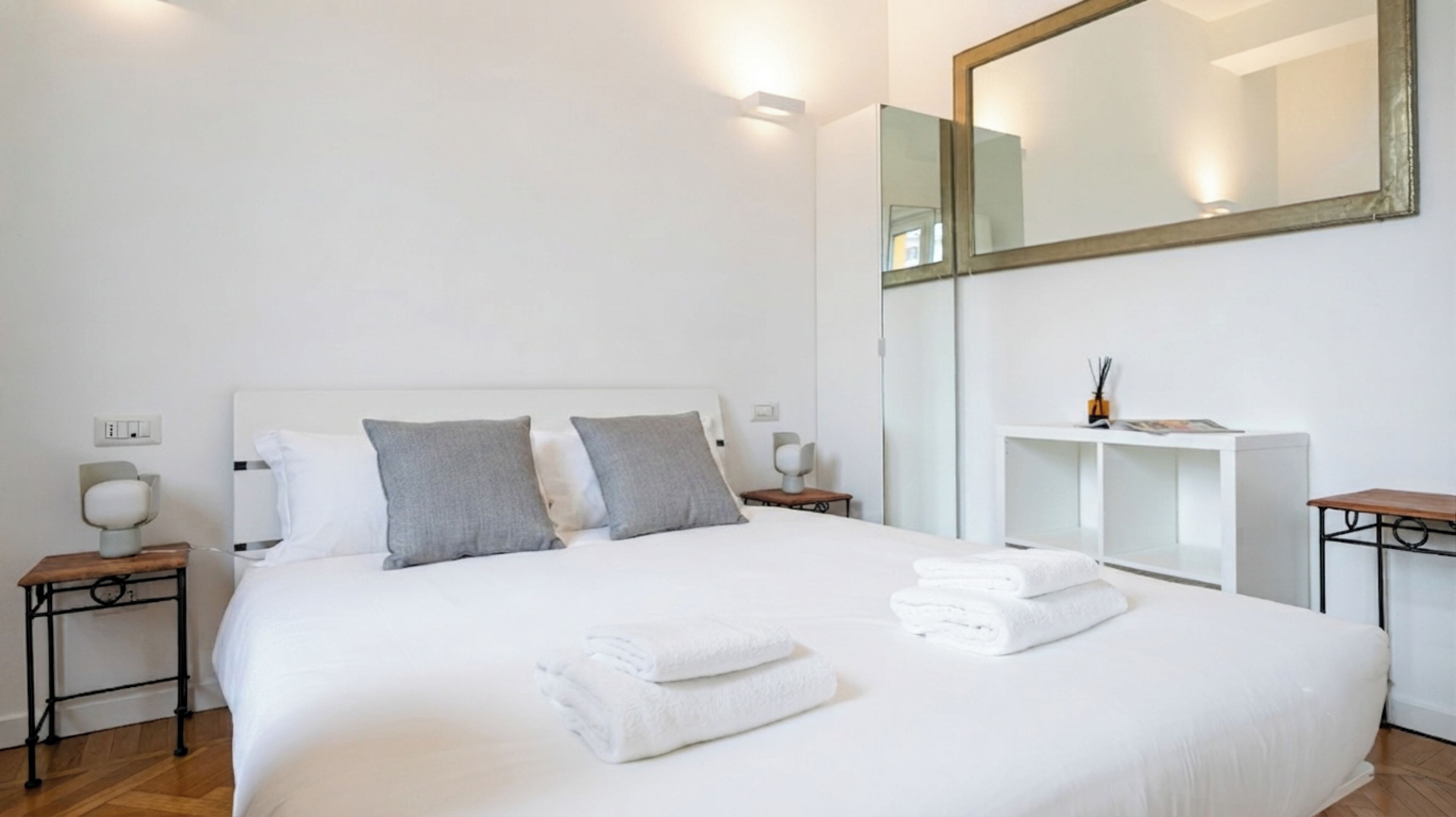 A hotel room with a large bed, a bedside table with a lamp, a mirror, and a small desk with a lamp.