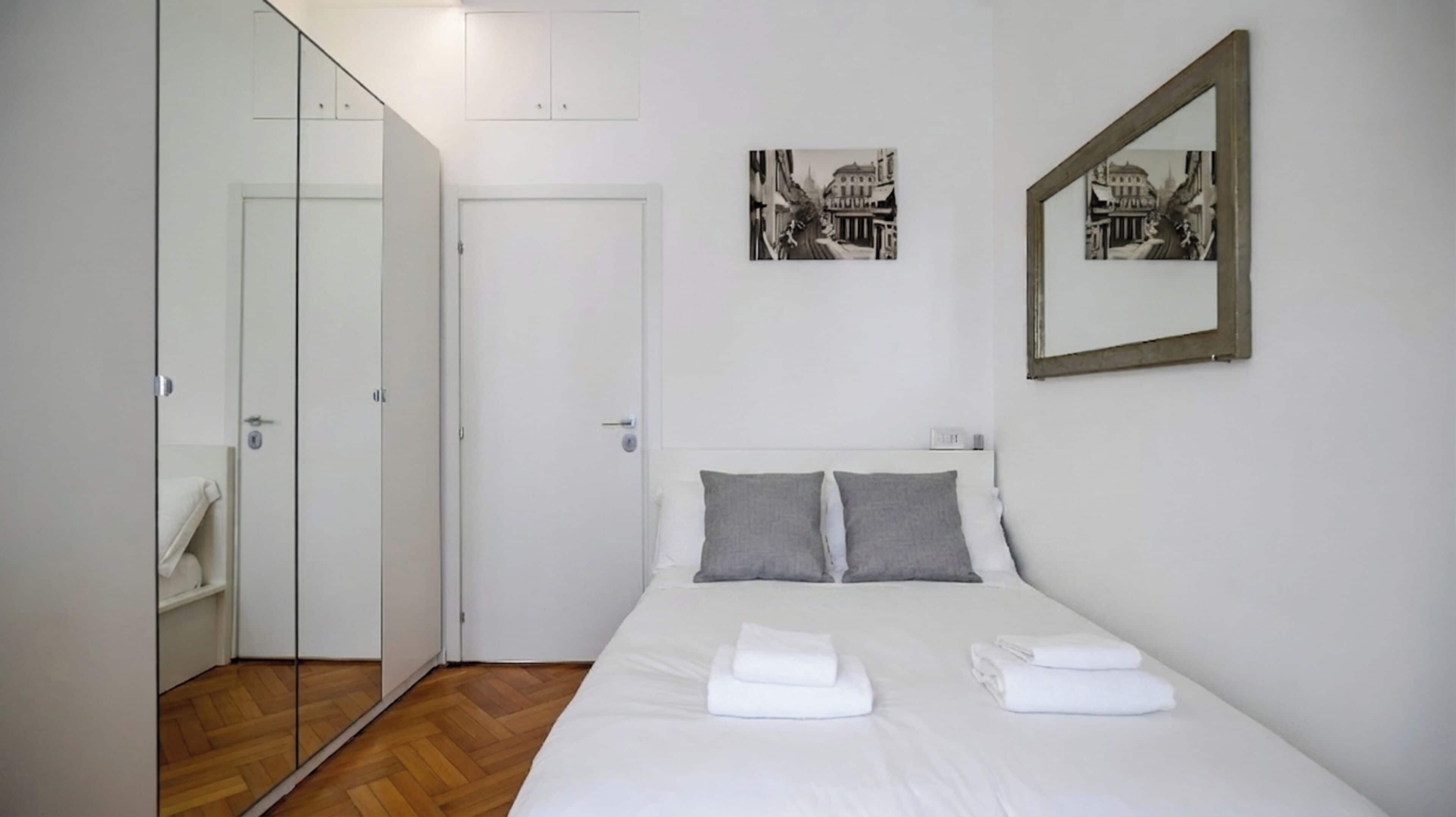 A bedroom with a bed, two pillows, a bedside table, a mirror, and a door with a towel hanging on it.