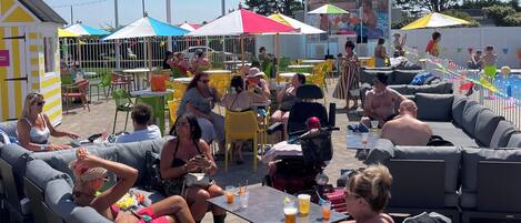 An outdoor seating area with grey sofas and tables, surrounded by colorful umbrellas and chairs.