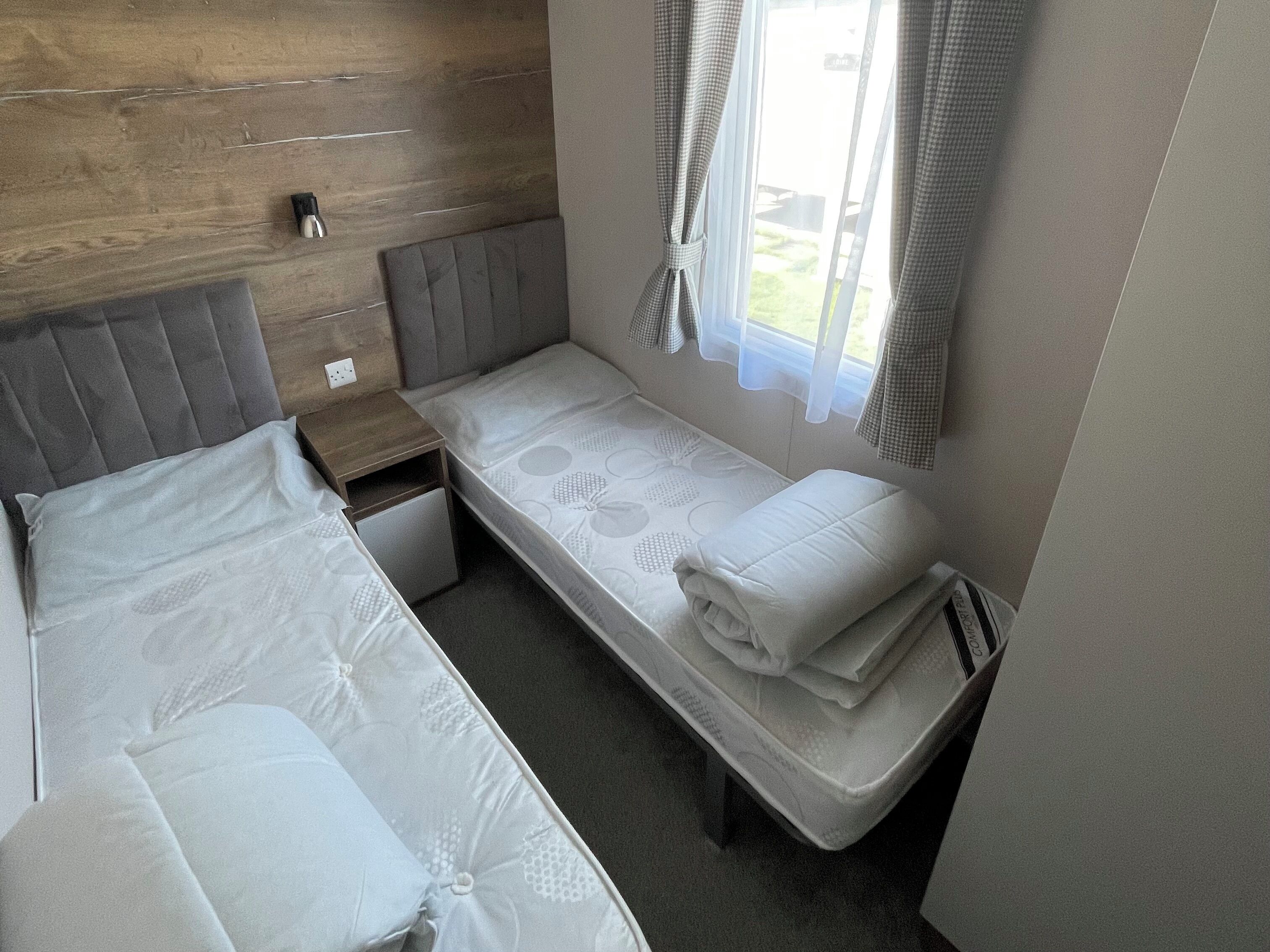 A compact room with two single beds, a small bedside table, a power outlet, and a window with curtains.