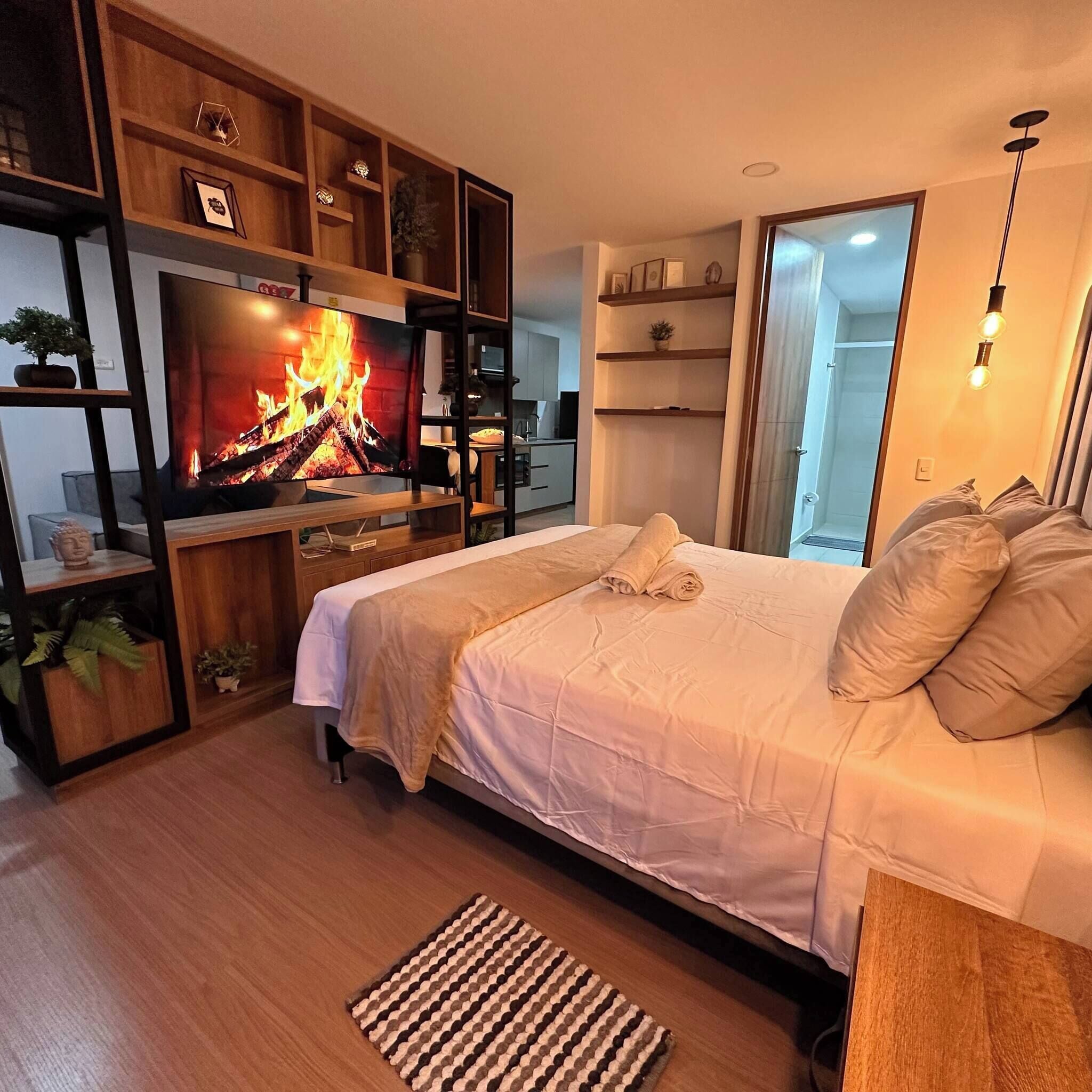 Une chambre moderne avec un téléviseur diffusant une image de cheminée, un lit avec des oreillers, un plancher en bois et un petit tapis.