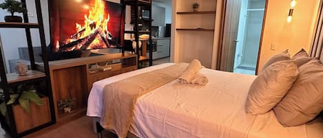 A modern bedroom with a TV displaying a fireplace, a bed with pillows, a wooden floor, and a small rug.