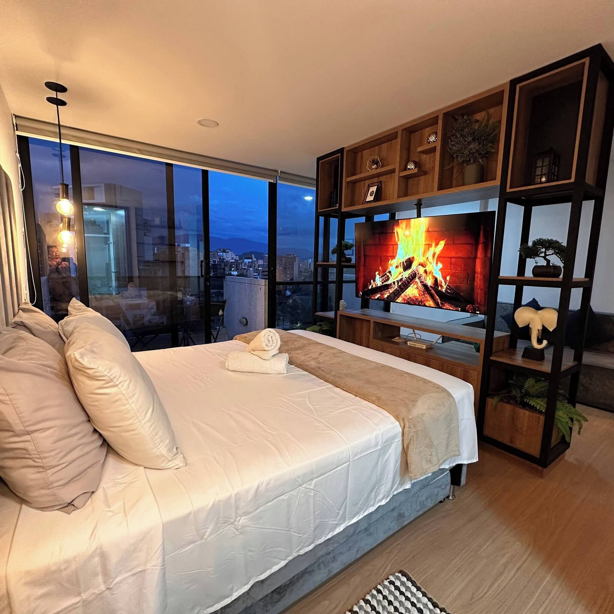 A modern bedroom with a large bed, a TV displaying a fireplace, and a city view through large windows.