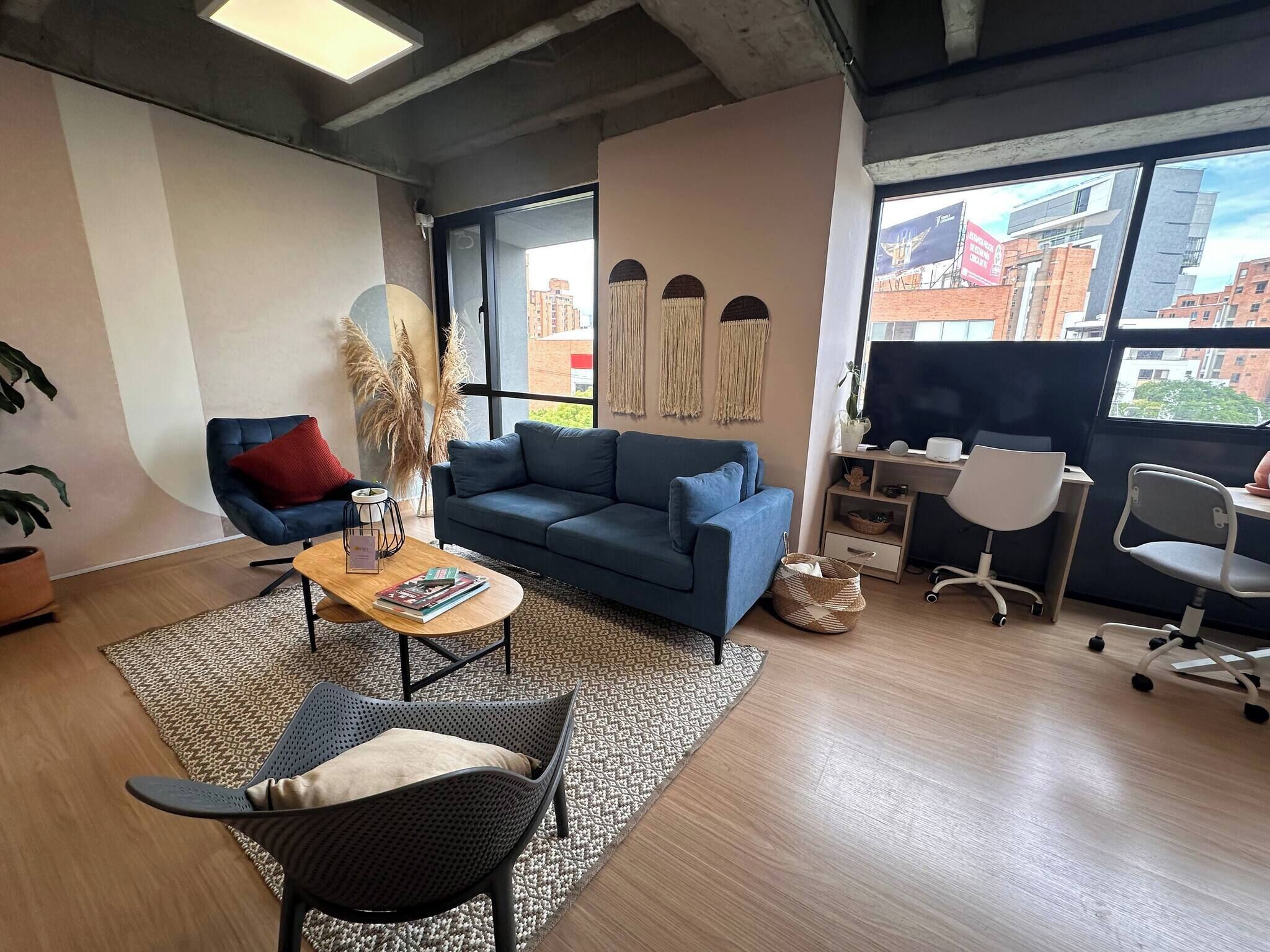 A modern living room with a blue sofa, a coffee table, and a chair. There is a large window with a view of buildings outside.
