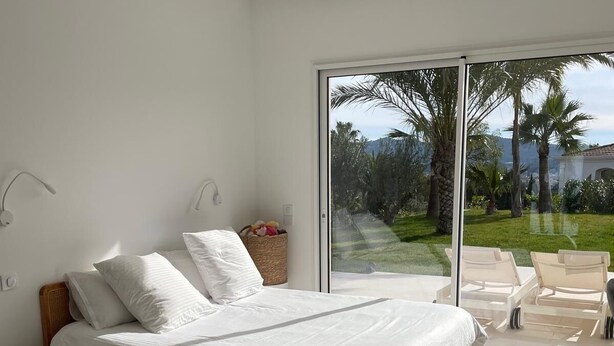 Ein modernes Schlafzimmer mit einem großen Bett, einem kleinen Nachttisch, einem Sessel und Blick auf einen Garten durch eine Schiebetür.