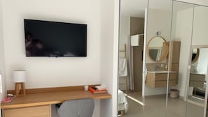 Ein modernes Schlafzimmer mit Schreibtisch aus Holz, einem grauen Stuhl, einem Bett mit weißer Bettwäsche, einem Fernseher und einem Schrank mit Spiegel.