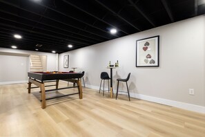 Ein Zimmer mit Billardtisch, zwei Stühlen, einem kleinen Tisch mit Flaschen und einem gerahmten Kunstwerk an der Wand.