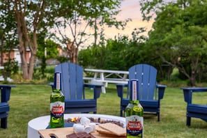 Zwei Flaschen Stella Artois Bier, eine Schokoladentafel und ein Marshmallow auf einem Tisch im Freien mit blauen Stühlen und einem Picknicktisch im Hintergrund.