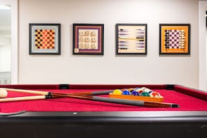 Ein Billardtisch mit Kugeln und Spielstangen, sowie vier gerahmte Brettspiele an der Wand.