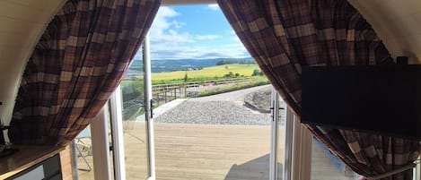 A room with a large arched window, wooden flooring, and a view of a landscape with fields and a distant hill.