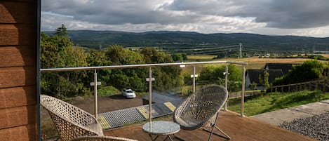 Ein Balkon mit Korbstühlen und einem Tisch, mit Blick auf eine ländliche Landschaft mit Hügeln und einem geparkten Auto im Hintergrund.