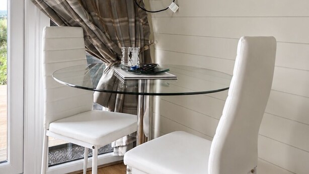 Un comedor con mesa de vidrio y sillas blancas, un televisor montado en la pared y un ventanal con cortinas.