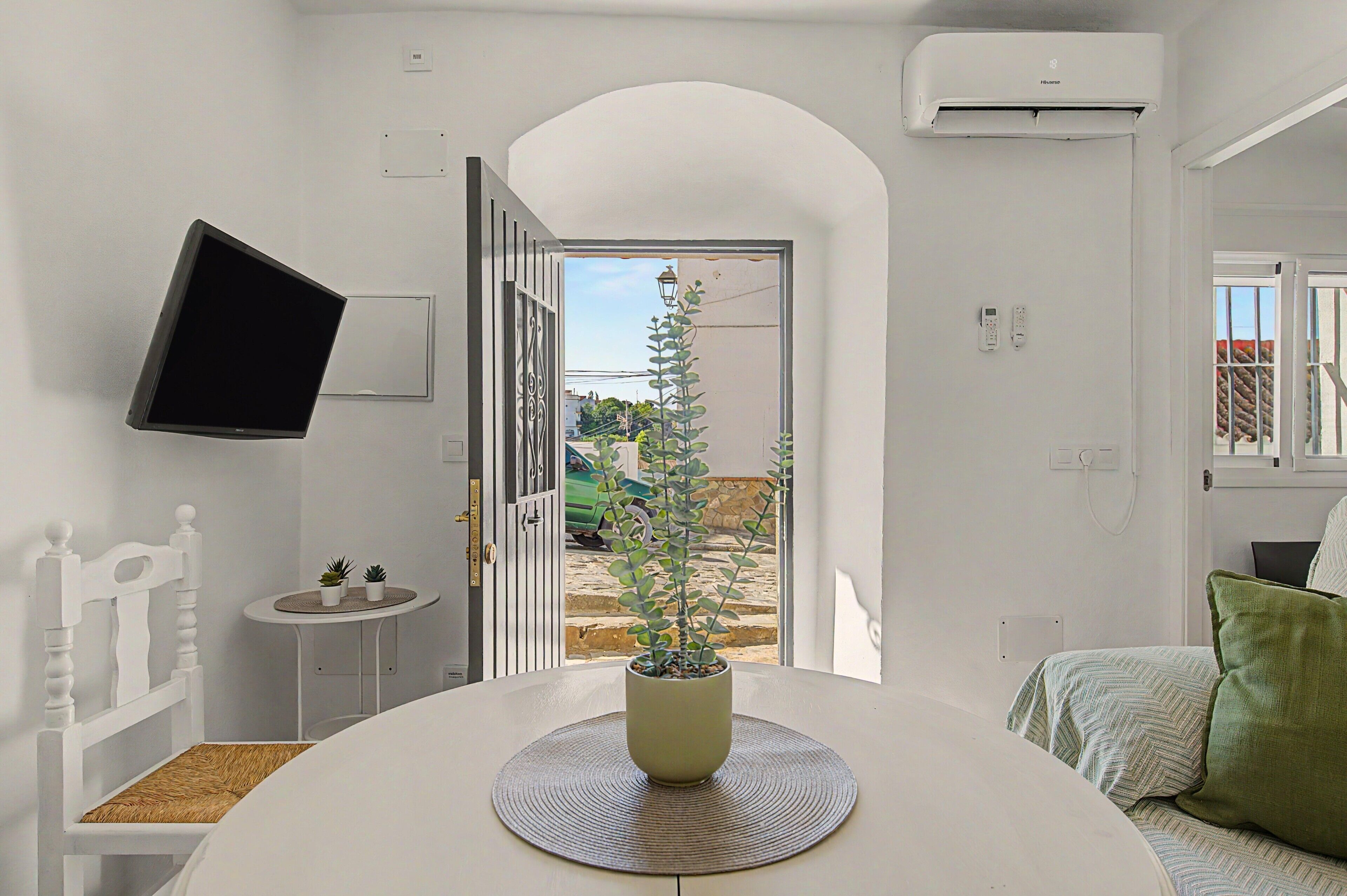 Een kamer met een eettafel, een aan de muur bevestigde televisie, een open deur naar buiten en een groene plant in het midden.