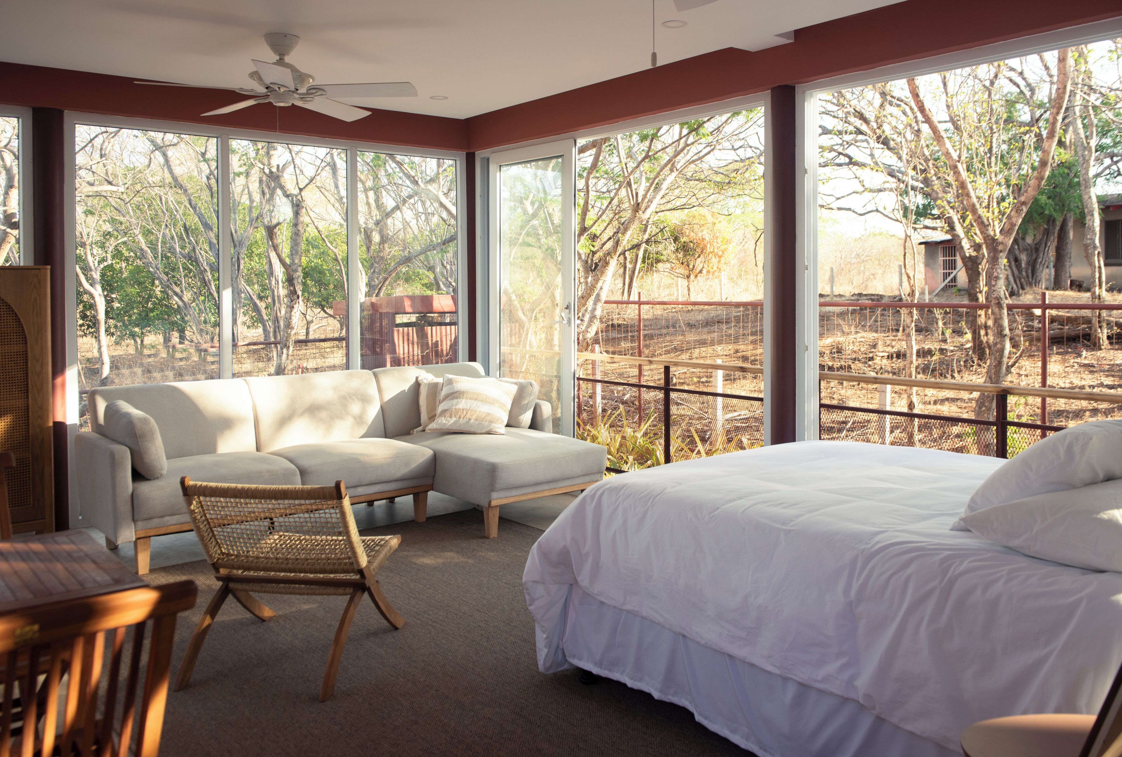 A bedroom with a bed, a sofa, and a chair. There are large windows with a view of trees and a fence.