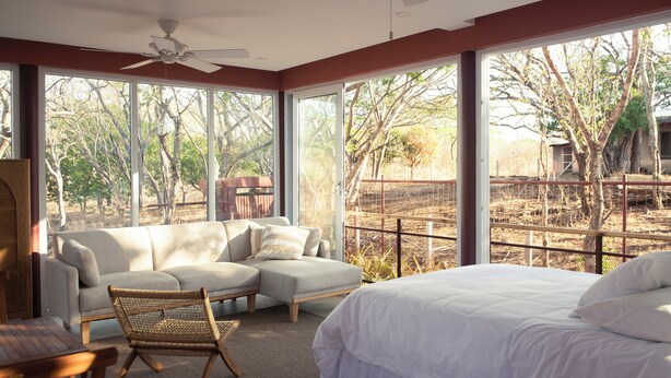 Quarto com cama, sofá e poltrona. Grandes janelas oferecem vista para árvores e uma cerca.