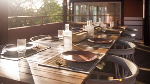Uma longa mesa de jantar posta com pratos, copos e talheres, com vista para árvores lá fora.