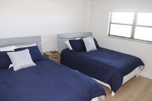 Ein Zimmer mit zwei Betten, jeweils mit Kopfteil und Kissen, einem Holz-Nachttisch und einem Fenster mit Jalousien.