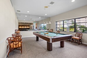 A pool table with surrounding chairs, a kitchen area in the background, and a view of a residential building through large windows. - Gulf and Bay Club - D506 (Siesta Key)