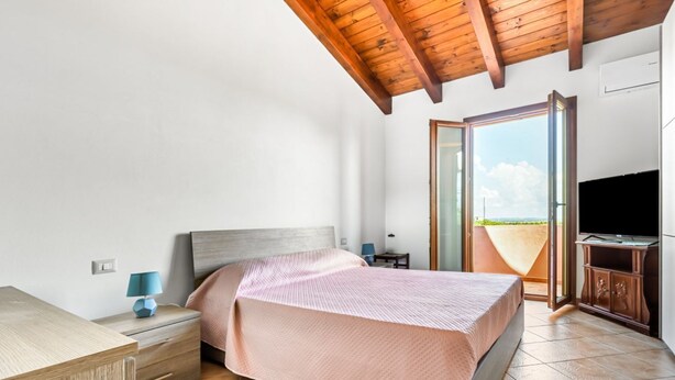 A bedroom with a bed, nightstand, lamp, TV, and a view of the outdoors through a glass door. - GuestHost - Sweet Home in Portoscuso! (Portoscuso)